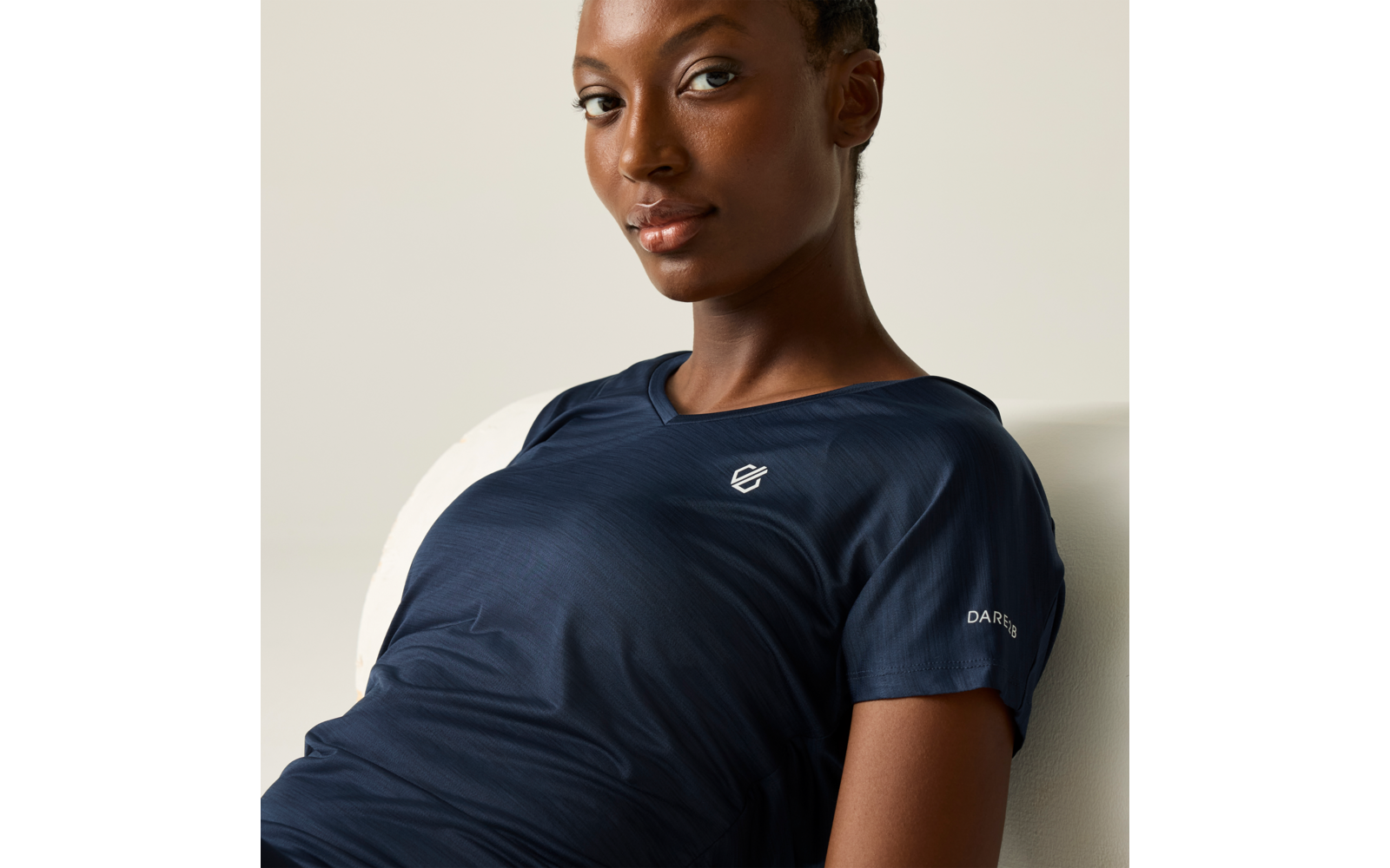 Person sitzt leicht zurückgelehnt auf hellem Stuhl, Nahaufnahme von Kopf und Oberkörper, trägt dunkelblaues T‑Shirt mit weißem Logo auf der Brust und kleinem Schriftzug am Ärmel, Blick zur Kamera