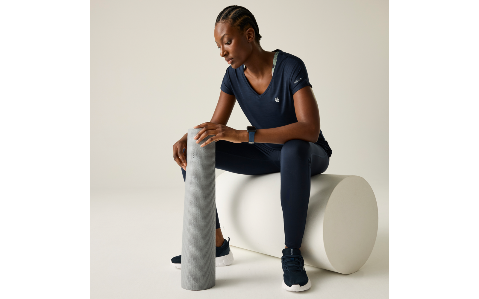 Person mit geflochtenem Haar in dunkelblauer Sportkleidung sitzt auf einem weißen Zylinder, hält eine graue Schaumstoffrolle senkrecht und blickt nach unten