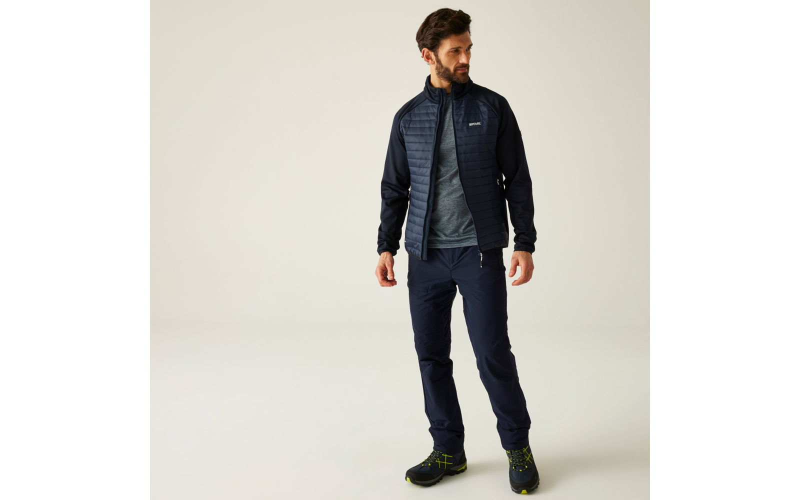 Mann mit Bart und dunklen Haaren steht vor hellem Hintergrund, trägt gesteppte dunkelblaue Jacke, graues T‑Shirt, dunkelblaue Hose und schwarze Wanderschuhe mit grünen Details; Blick nach rechts