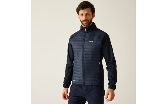 Person mit dunklem Haar und kurzem Bart, trägt gesteppte dunkelblaue Jacke mit Reißverschluss und dunkle Hose, vor hellem Hintergrund