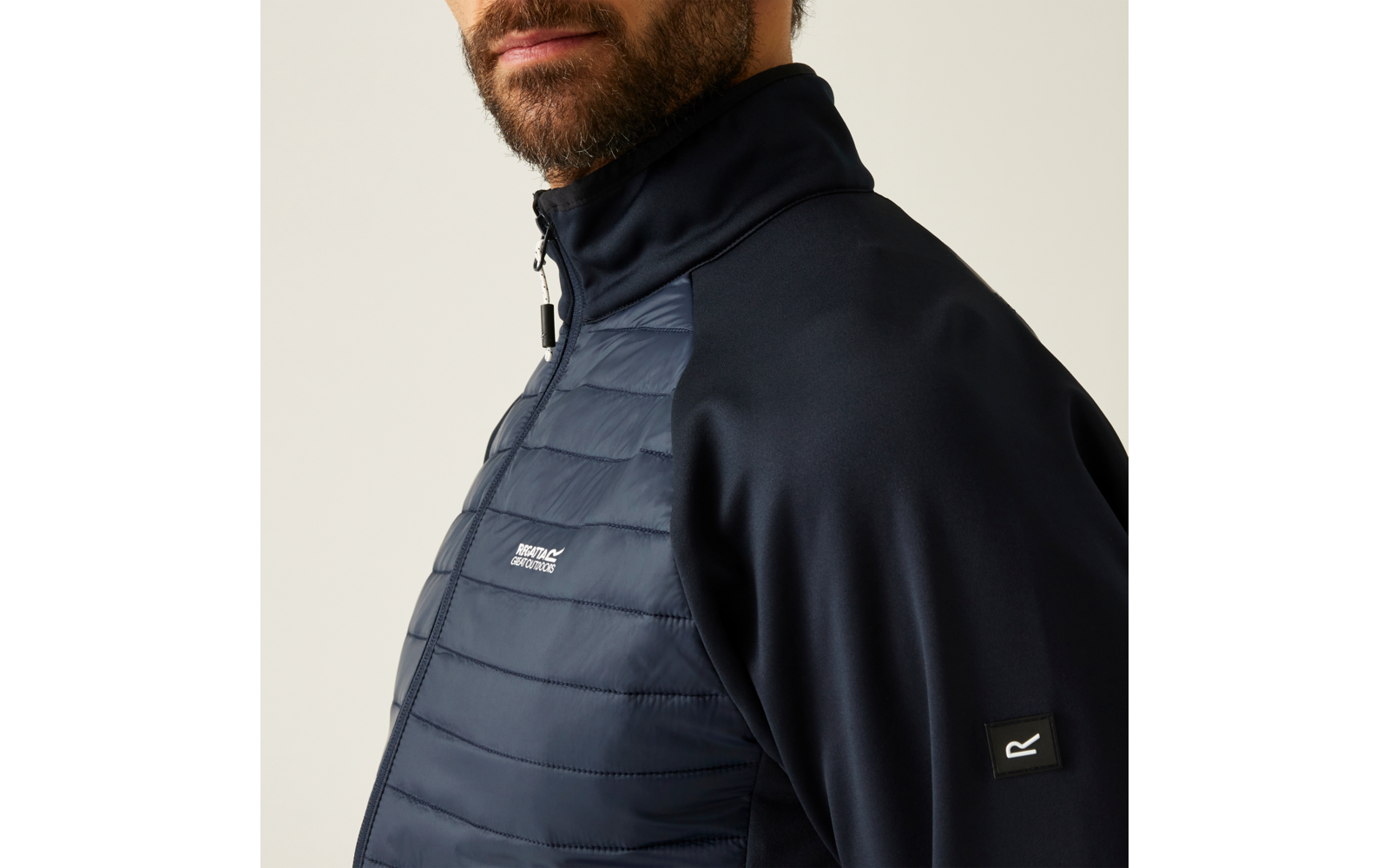 Person von Brust bis Kinn in dunkelblauer Jacke mit gesteppter Vorderseite, Reißverschluss, hohem Kragen, sichtbarem Bart und kleinem Logo-Patch am Ärmel