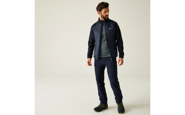 Mann mit Bart und dunklen Haaren steht vor hellem Hintergrund, trägt gesteppte dunkelblaue Jacke, graues T‑Shirt, dunkelblaue Hose und schwarze Wanderschuhe mit grünen Details; Blick nach rechts