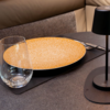Oranger Teller mit weißem Sprenkelmuster auf dunkler Tischunterlage, links ein klares stieloses Glas, rechts Besteck und eine schwarze Tischlampe; beiger Polstersitz im Hintergrund