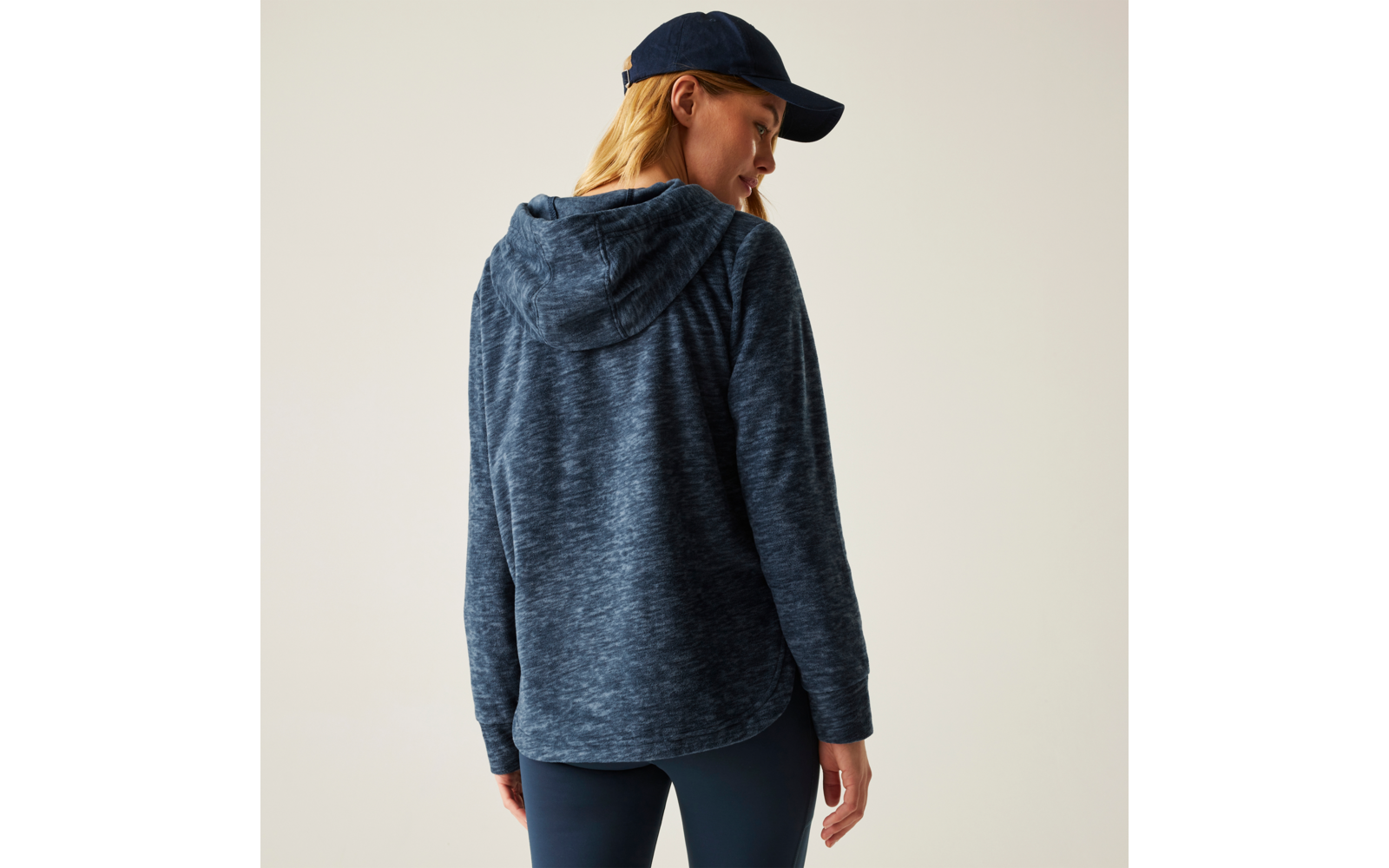 Person von hinten, trägt dunkelblauen Kapuzenpullover, dunkelblaue Baseballkappe und dunkle Leggings, Kopf leicht zur Seite gedreht