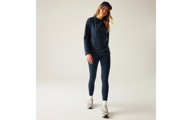 Frau steht in dunkelblauem Hoodie, dunkelblauen Leggings und dunkelblauer Baseballmütze, trägt weiße Turnschuhe vor neutralem Hintergrund