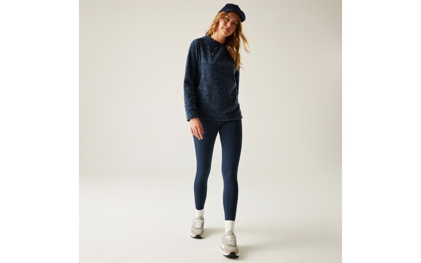 Frau steht in dunkelblauem Hoodie, dunkelblauen Leggings und dunkelblauer Baseballmütze, trägt weiße Turnschuhe vor neutralem Hintergrund
