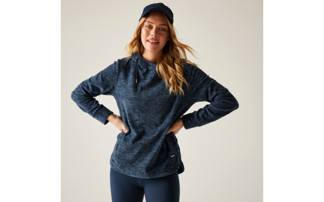 Frau vor hellem Hintergrund, trägt dunkelblaues Cap, dunkelblauen Kapuzenpullover und dunkle Leggings, Hände auf den Hüften, lächelnd