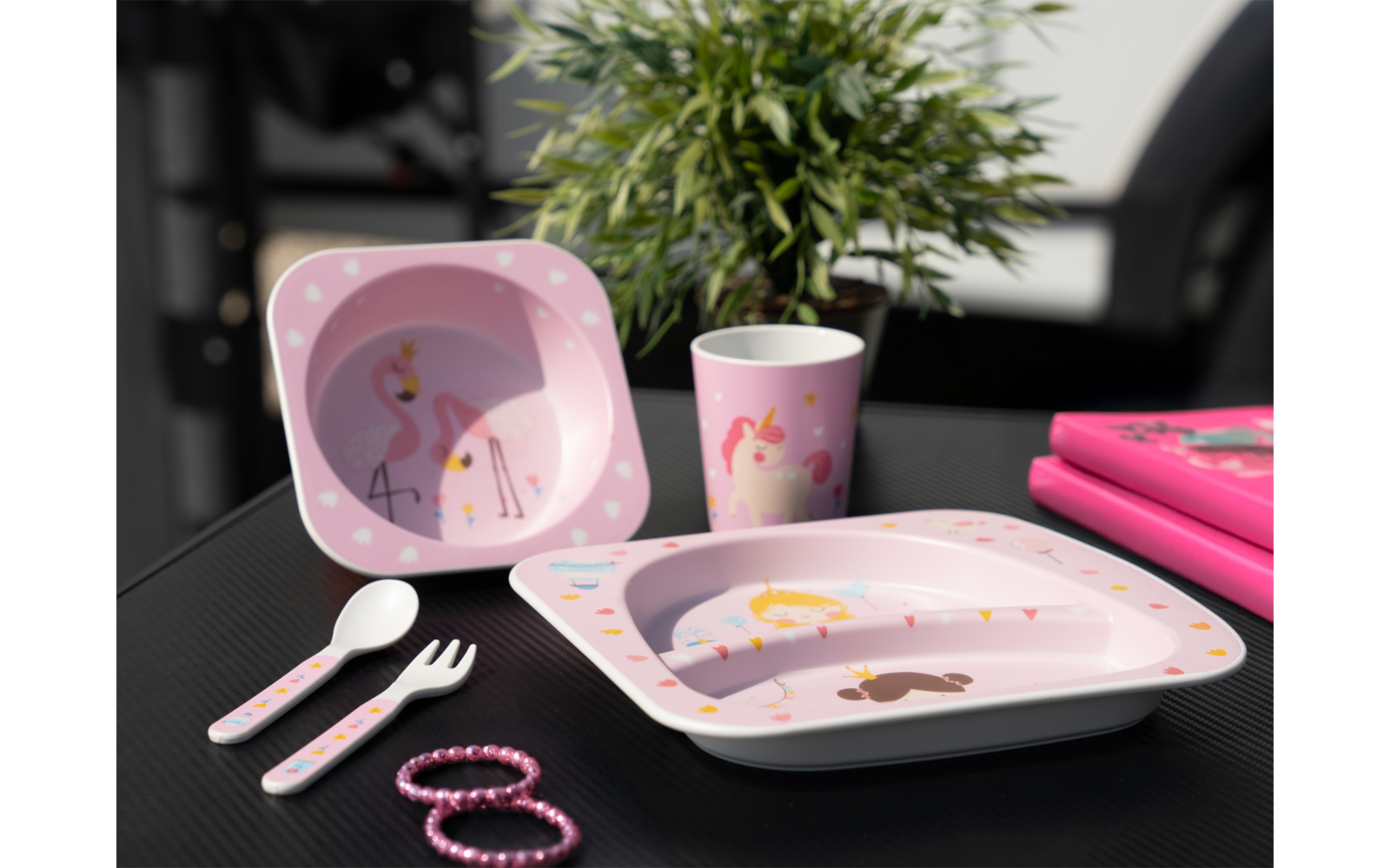 Kindergeschirr in Rosa mit Flamingo-Motiv, bestehend aus Teller, Schale, Becher, Besteck und Armbändern auf einem schwarzen Tisch, im Hintergrund eine grüne Pflanze