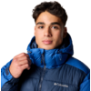 Porträt einer Person mit dunklen, kurzen, gewellten Haaren, die eine blaue Columbia-Steppjacke mit hochgezogenem Kragen trägt; rechte Hand hält den Reißverschluss; weißer Hintergrund