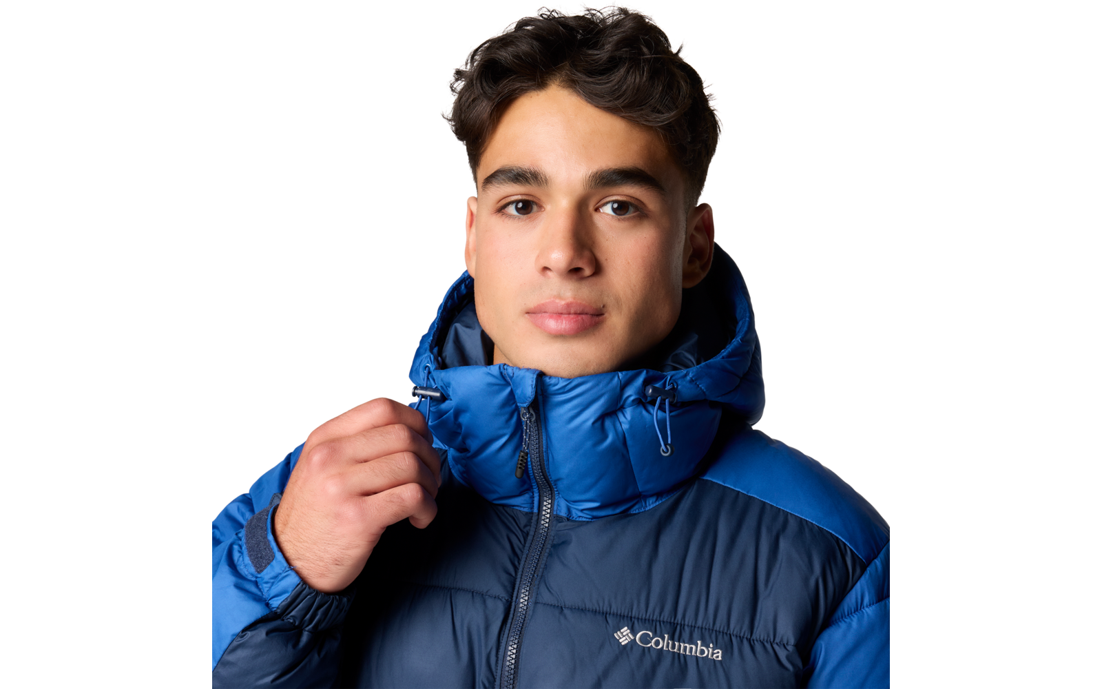 Porträt einer Person mit dunklen, kurzen, gewellten Haaren, die eine blaue Columbia-Steppjacke mit hochgezogenem Kragen trägt; rechte Hand hält den Reißverschluss; weißer Hintergrund