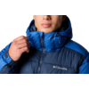 Porträt einer Person mit dunklen, kurzen, gewellten Haaren, die eine blaue Columbia-Steppjacke mit hochgezogenem Kragen trägt; rechte Hand hält den Reißverschluss; weißer Hintergrund