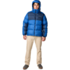 Person steht frontal, trägt blaue Steppjacke mit Kapuze, beige Hose und dunkle Schuhe, heller Hintergrund