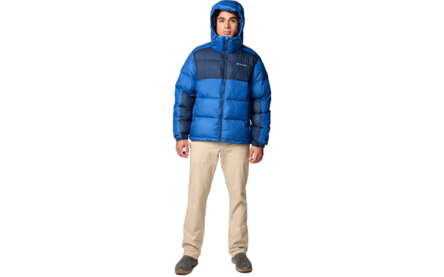 Person steht frontal, trägt blaue Steppjacke mit Kapuze, beige Hose und dunkle Schuhe, heller Hintergrund
