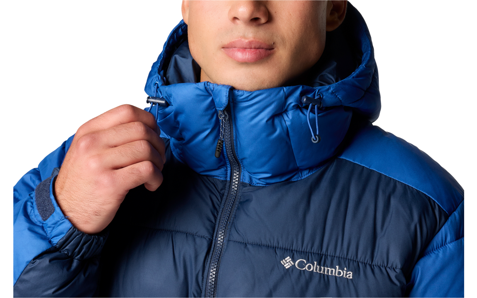 Porträt einer Person mit dunklen, kurzen, gewellten Haaren, die eine blaue Columbia-Steppjacke mit hochgezogenem Kragen trägt; rechte Hand hält den Reißverschluss; weißer Hintergrund