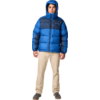Person steht frontal, trägt blaue Steppjacke mit Kapuze, beige Hose und dunkle Schuhe, heller Hintergrund