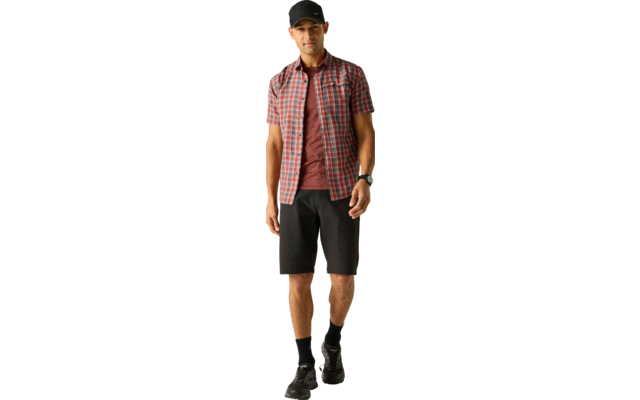 Person mit schwarzer Kappe, rotem T‑Shirt, rot kariertem Kurzarmhemd, schwarzen Shorts, schwarzen Socken, schwarzen Schuhen und Smartwatch am Handgelenk vor hellem einfarbigem Hintergrund
