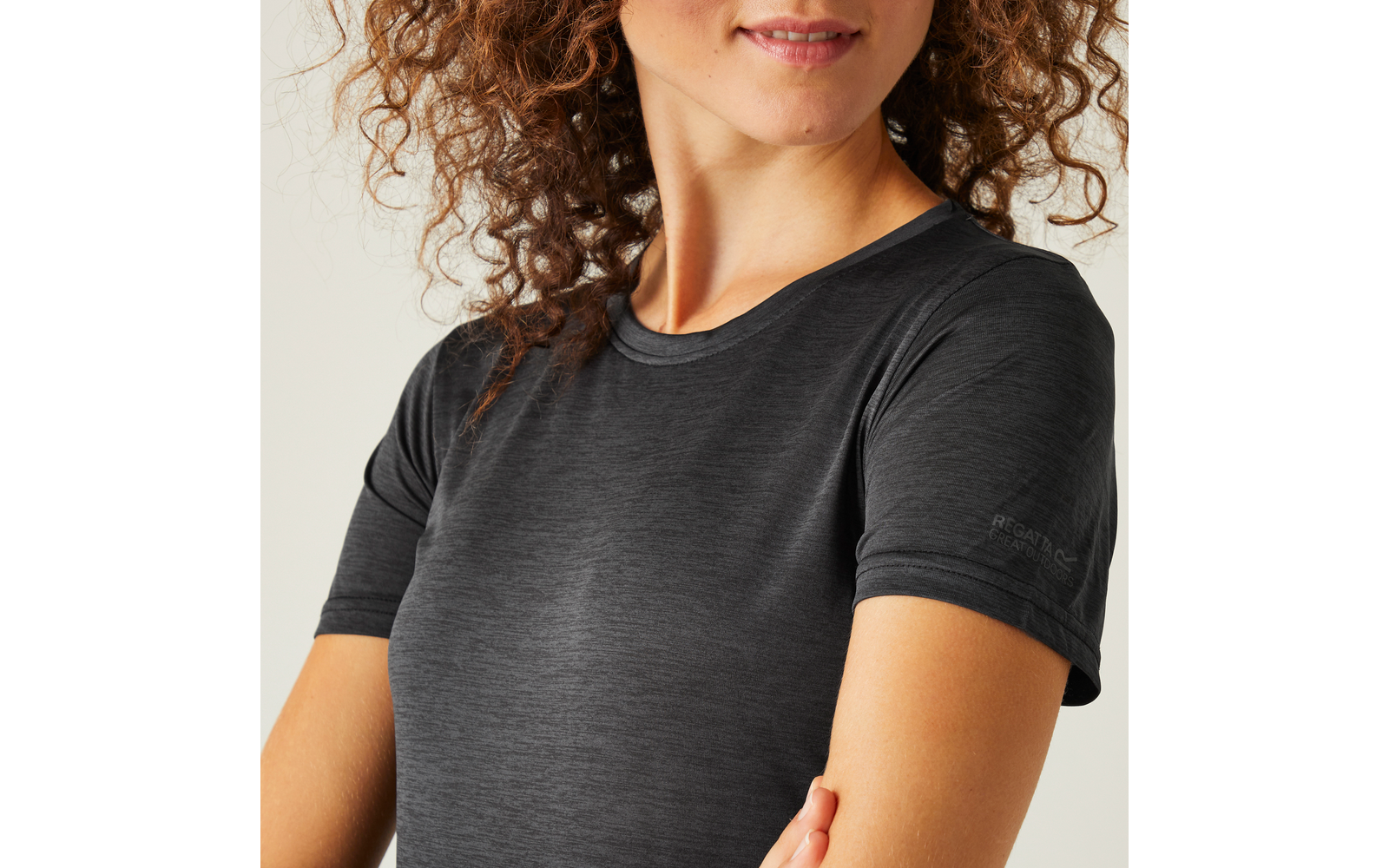 Teilansicht einer Person von Brust bis Kinn, lockiges braunes Haar, dunkelgraues kurzärmeliges T-Shirt, Arme gekreuzt, leichtes Lächeln