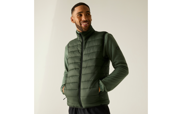 Homme portant une veste matelassée vert foncé sur un haut vert à manches longues, mains dans les poches, souriant, fond uni clair