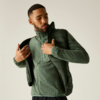 Homme en train d'enfiler une doudoune sans manches olive sur un zip polaire vert; fond uni clair