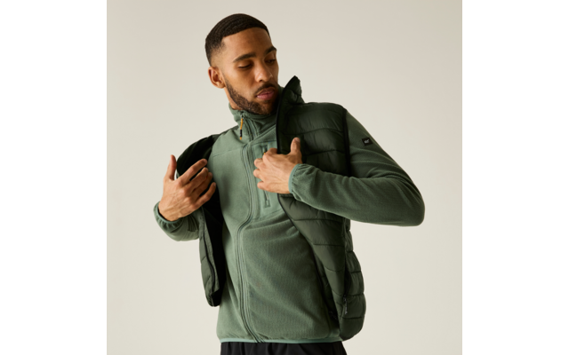 Homme en train d'enfiler une doudoune sans manches olive sur un zip polaire vert; fond uni clair