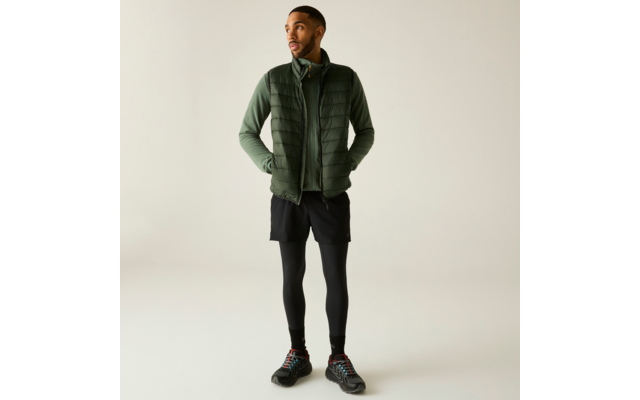 Personne debout portant une doudoune sans manches verte sur un haut à manches longues vert, shorts noirs sur des leggings noirs et chaussures de sport noires, mains dans les poches, fond clair uni