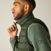 Profil latéral d'un homme aux cheveux courts et à la barbe tenant le col d'une doudoune sans manches vert foncé; il porte un vêtement vert texturé à manches longues sur un fond clair et neutre