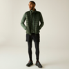 Personne debout portant une doudoune sans manches verte sur un haut à manches longues vert, shorts noirs sur des leggings noirs et chaussures de sport noires, mains dans les poches, fond clair uni