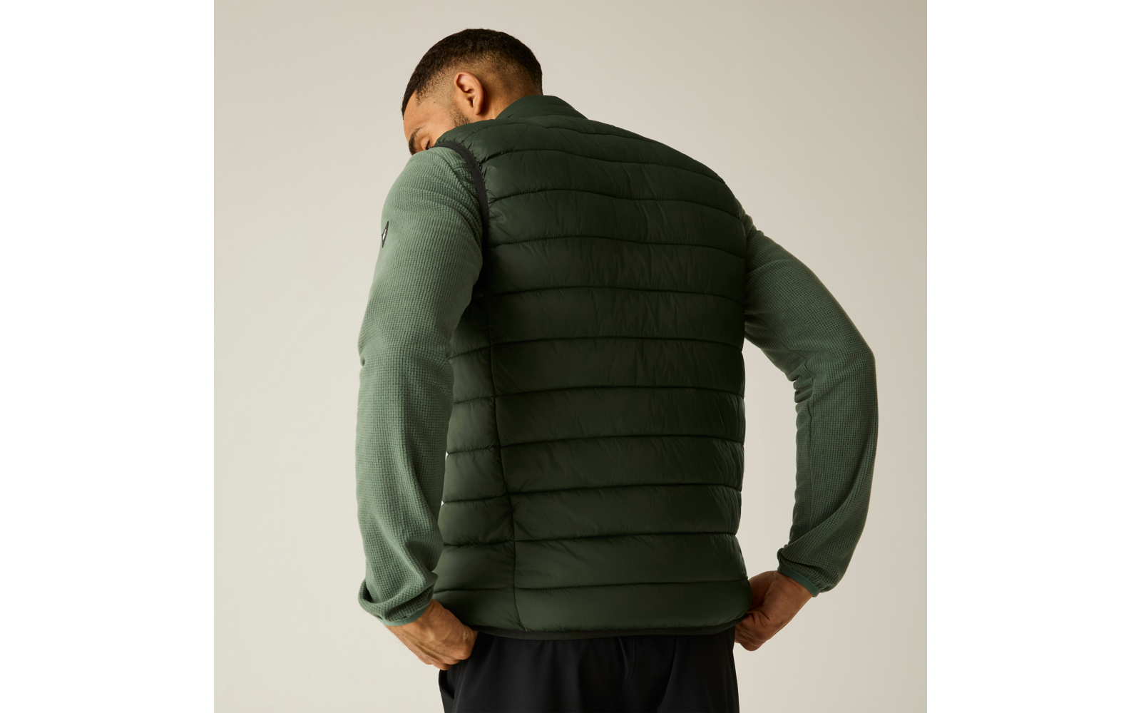 Personne vue de dos portant une doudoune sans manches matelassée vert foncé sur un haut manches longues vert clair, mains sur les hanches, pantalon noir, fond uni clair