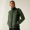Homme portant une veste matelassée vert foncé sur un haut vert à manches longues, mains dans les poches, souriant, fond uni clair