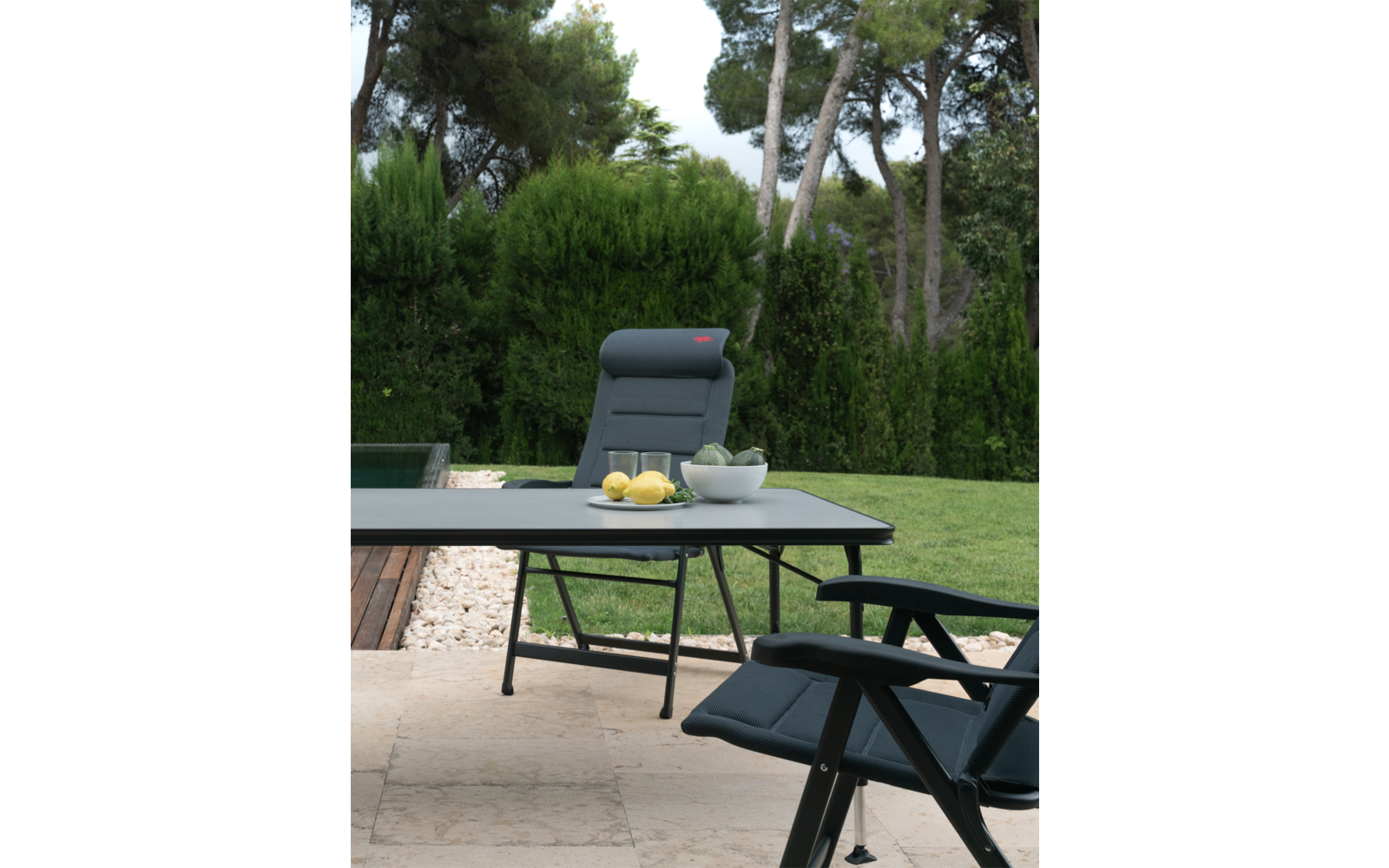 Ein grüner Garten mit einem schwarzen Tisch und zwei schwarzen Stühlen, auf dem Tisch steht eine Schale mit Zitronen