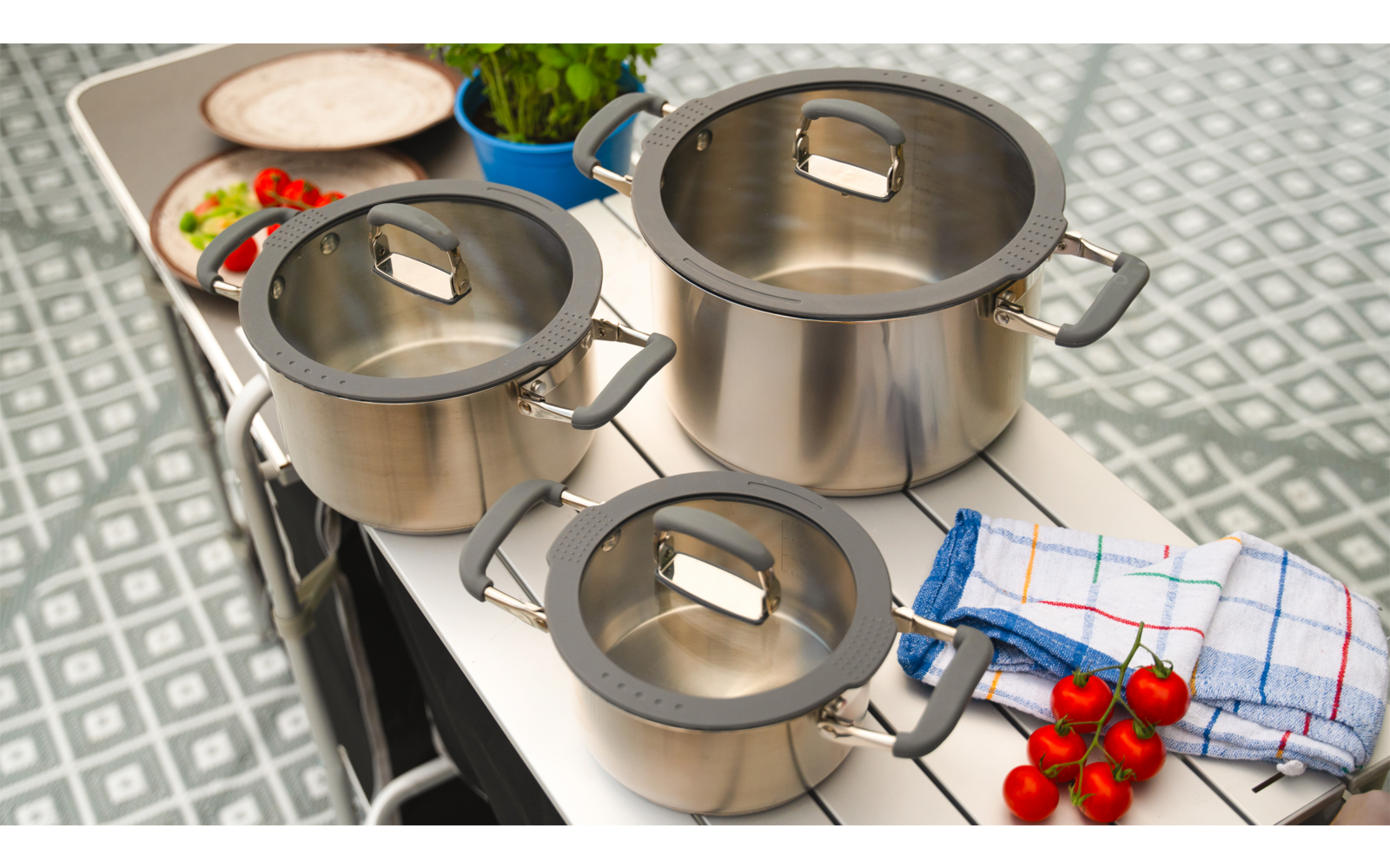 Küchenarbeitsfläche mit drei Edelstahlkochtöpfen, einem Schneidebrett mit Gemüse und einem roten Küchenwerkzeug