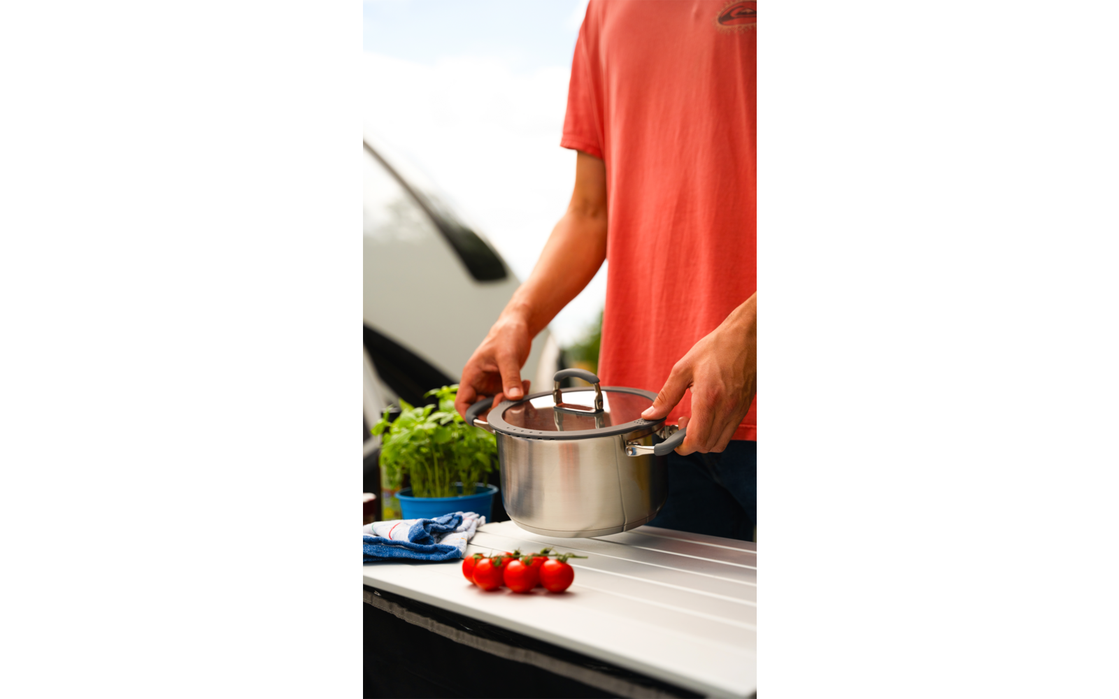 Person in rotem T-Shirt kocht mit Topf und Schneebesen in der Küche, drei Tomaten und frische Kräuter auf der Arbeitsplatte