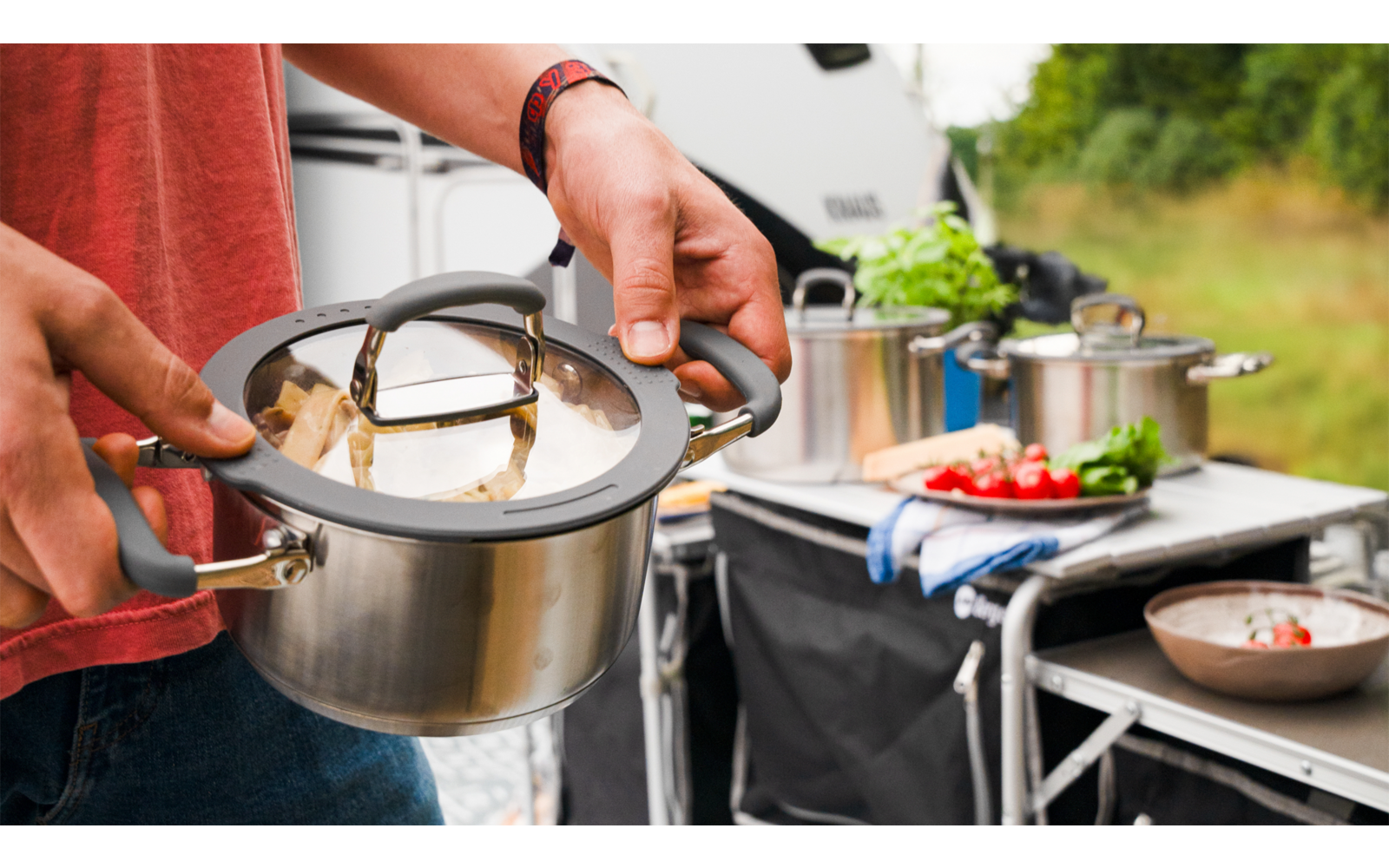 Person gießt Wasser aus einem kleinen Kessel in einen größeren Topf im Freien