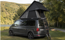 Grauer Camper-Van mit aufgestelltem Dachzelt und Leiter auf einer Wiese neben einem See, Bäume und bewaldete Hügel im Hintergrund