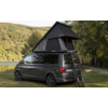 Grauer Camper-Van mit aufgestelltem Dachzelt und Leiter auf einer Wiese neben einem See, Bäume und bewaldete Hügel im Hintergrund
