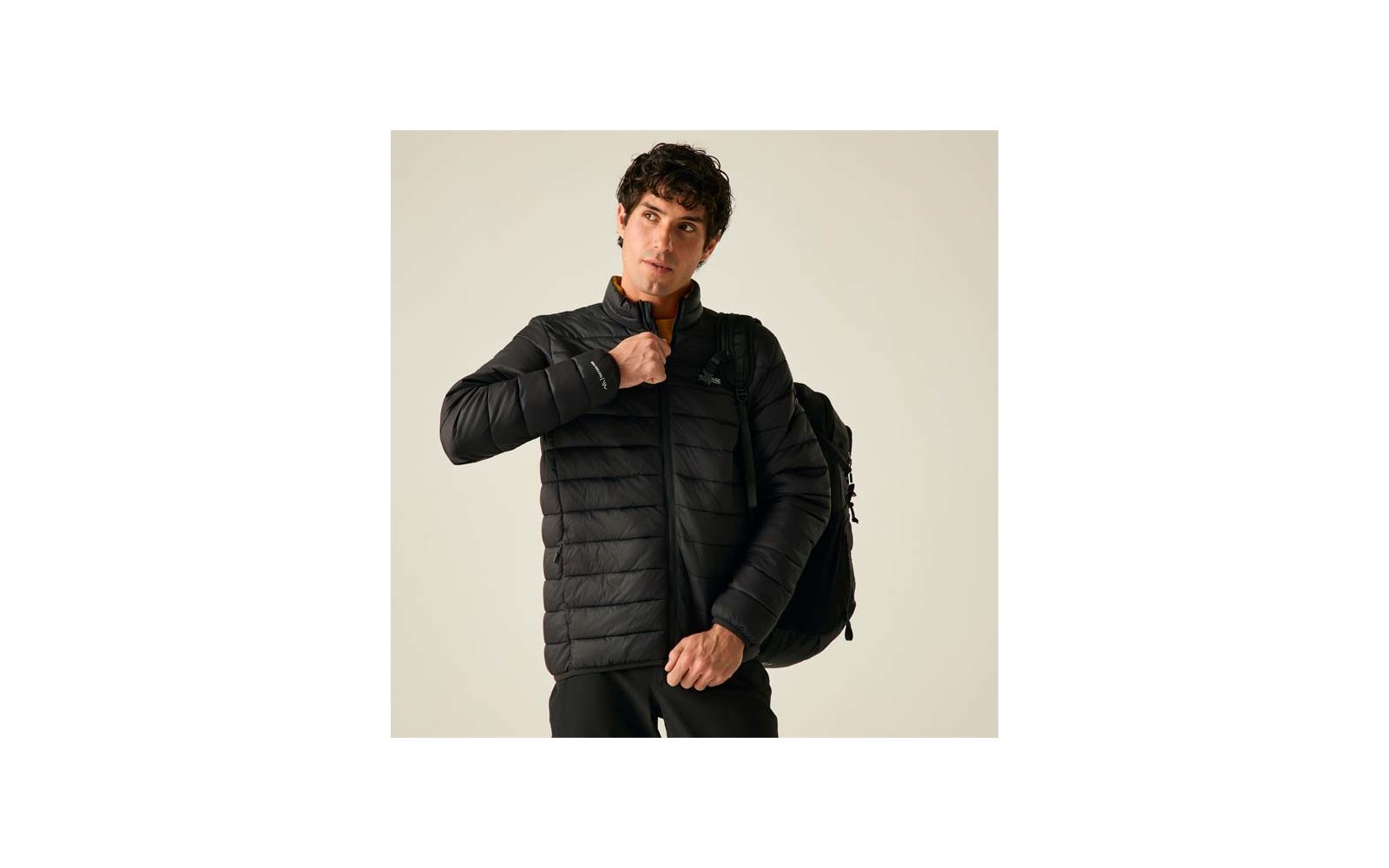 Ein Mann mit dunklen Haaren trägt eine schwarze gesteppte Jacke und einen Rucksack, er hält den Reißverschluss und blickt nach rechts vor hellem Hintergrund
