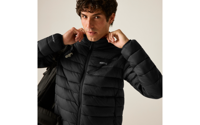 Mann mit dunklem, lockigem Haar zieht den Kragen einer schwarzen Steppjacke hoch, Schulterriemen einer Tasche sichtbar, neutraler Hintergrund