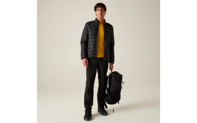 Stehende Person, schwarze gesteppte Jacke, gelbes Sweatshirt, schwarze Hose und Schuhe, hält schwarzen Rucksack in einer Hand, heller neutraler Hintergrund