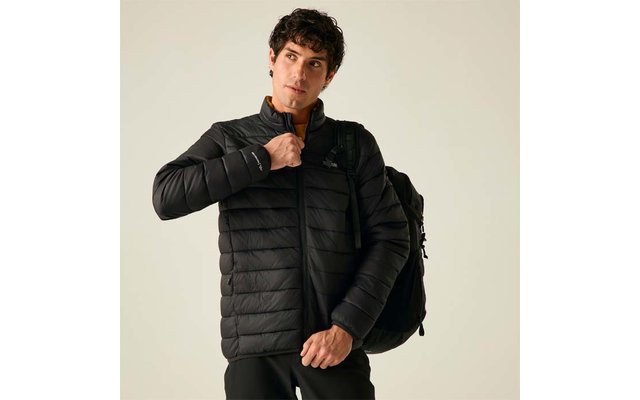 Ein Mann mit dunklen Haaren trägt eine schwarze gesteppte Jacke und einen Rucksack, er hält den Reißverschluss und blickt nach rechts vor hellem Hintergrund