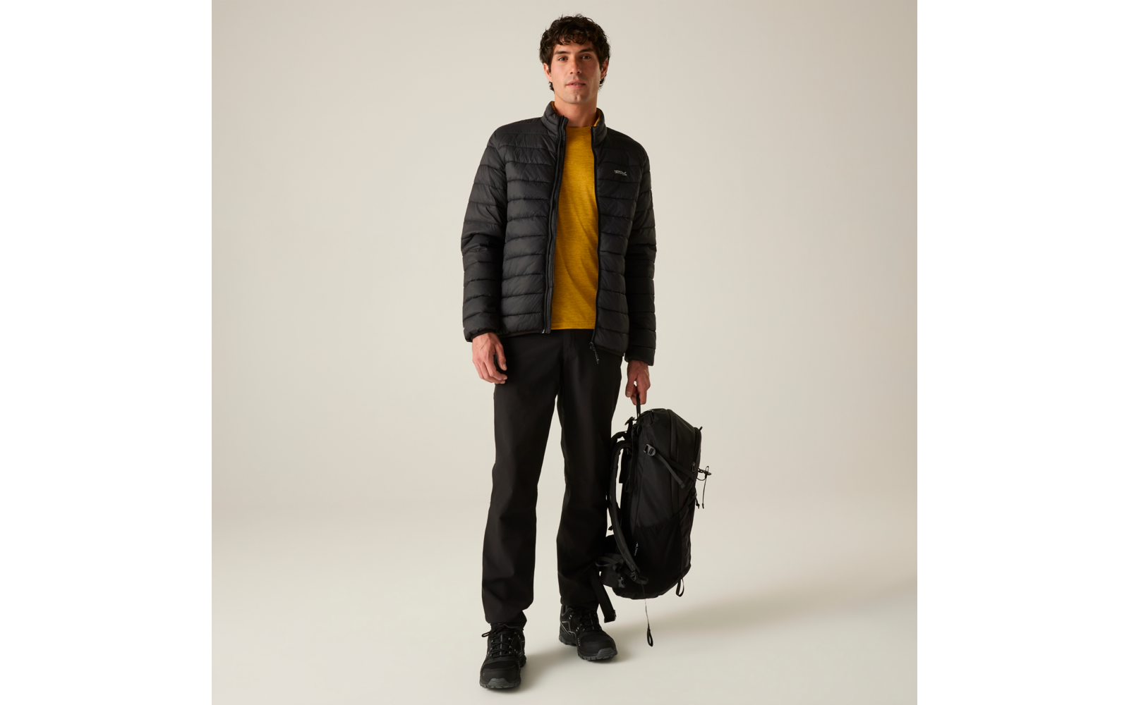 Stehende Person, schwarze gesteppte Jacke, gelbes Sweatshirt, schwarze Hose und Schuhe, hält schwarzen Rucksack in einer Hand, heller neutraler Hintergrund