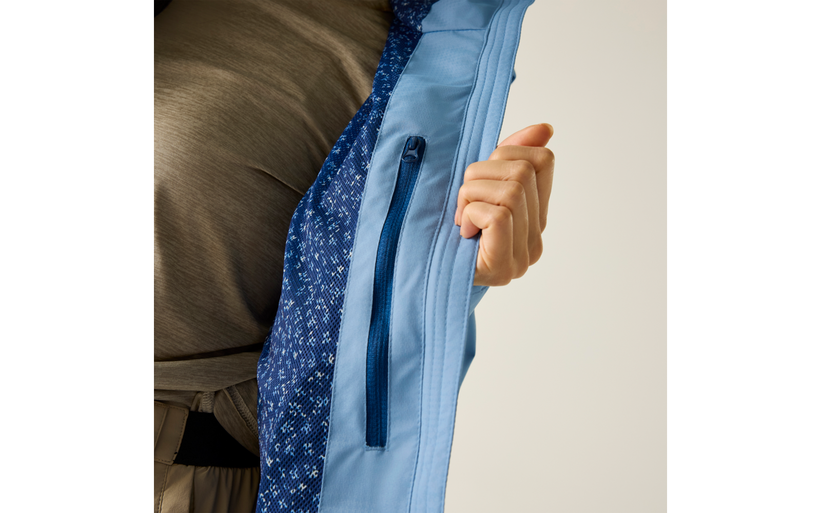 Hand hält die aufgeklappte Innenseite einer hellblauen Jacke mit sichtbarer Reißverschluss-Innentasche und dunkelblauem, gemustertem Futter; darunter ist ein graues Shirt sichtbar