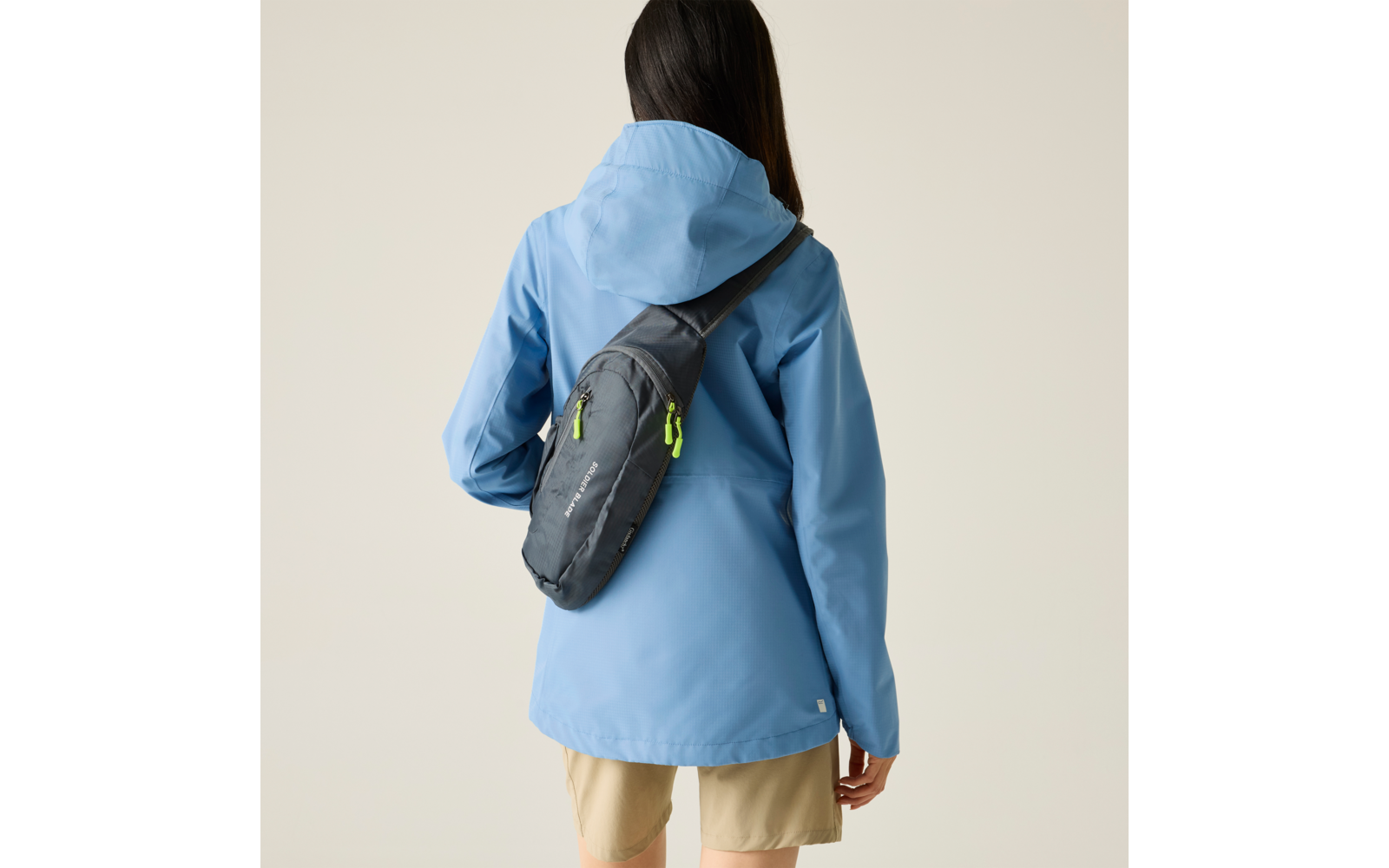 Person von hinten, trägt hellblaue Kapuzenjacke, beige Shorts und graue Umhängetasche quer über dem Rücken mit neongrünen Reißverschlüssen