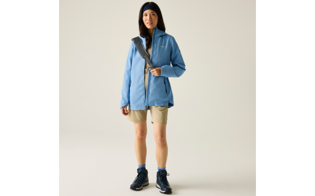 Person steht frontal vor hellem Hintergrund, trägt hellblaue Jacke, beige Shorts, blaue Socken, dunkle geschnürte Schuhe, schwarzes Stirnband und eine Umhängetasche