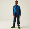 Een jongen staat voor een beige muur, draagt een blauwe jas, donkere broek en groene schoenen