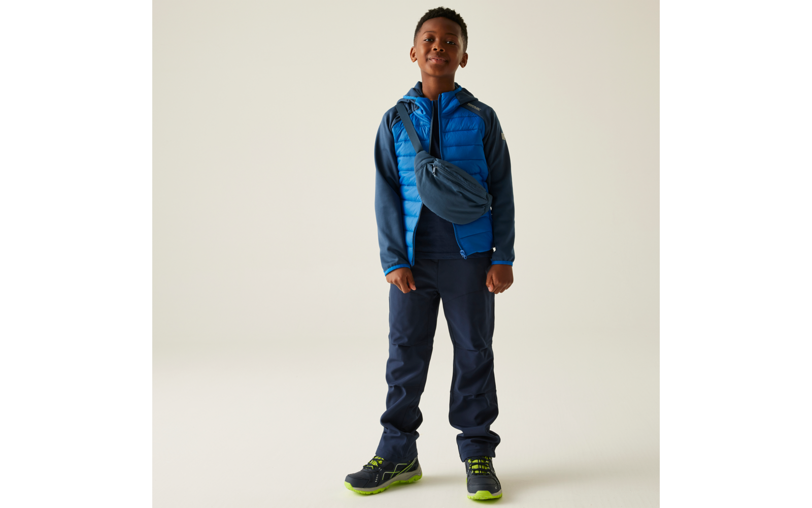 Een jongen staat voor een beige muur, draagt een blauwe jas, donkere broek en groene schoenen