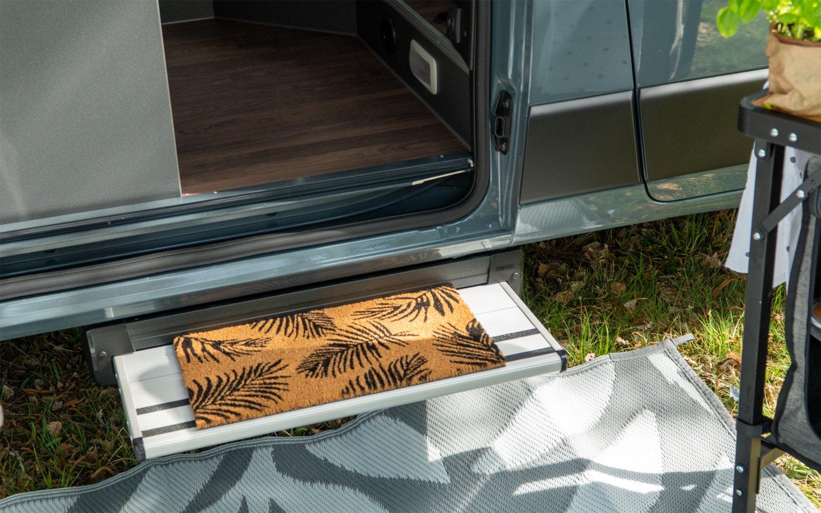 Porte coulissante de van ouverte, marche métallique avec paillasson brun à motif de fougères noires, tapis gris-blanc, herbe et partie d'une petite table avec plante