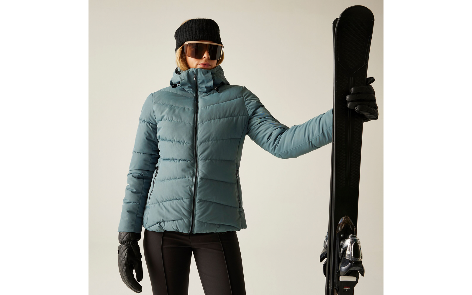 Frau in blauer Winterjacke, schwarzer Mütze und Handschuhen hält einen Ski