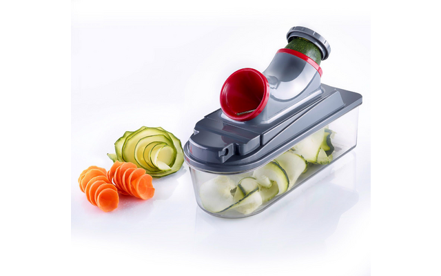 Gray device with red funnel on a clear plastic container holding thin zucchini ribbons; to the left spiral carrot slices and a zucchini curl on a white background