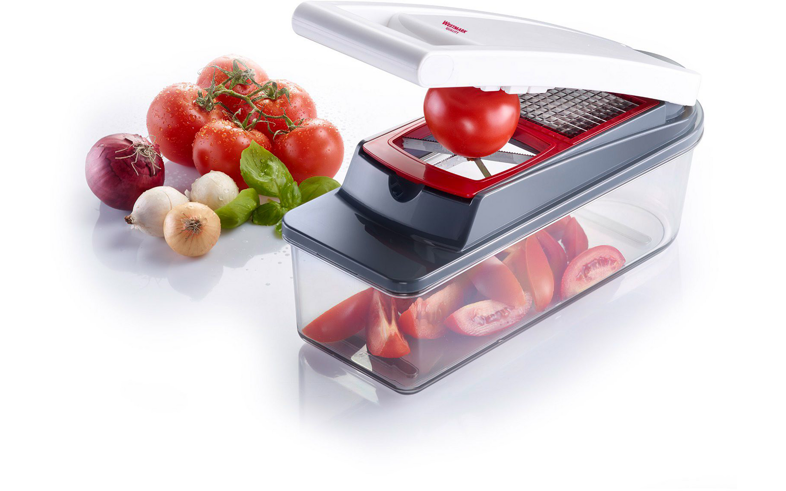White and gray plastic slicer with transparent container; a tomato is being pressed through a grid blade; in the background whole tomatoes, a red onion, a small yellow onion and basil leaves