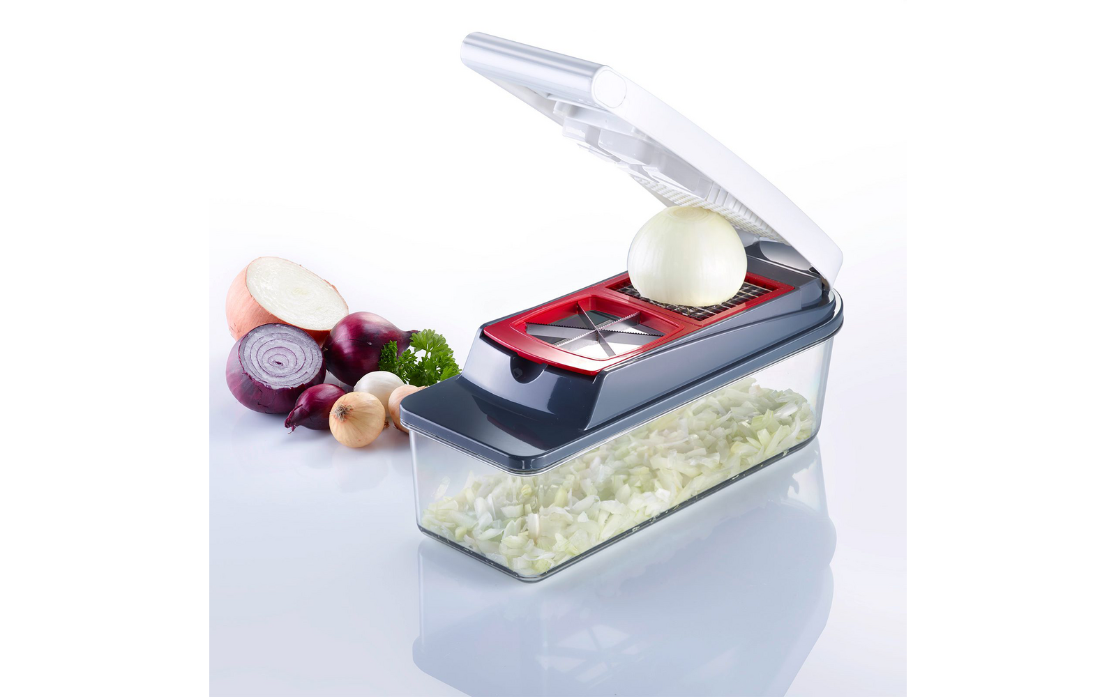 Plastic device with open white lid, red blade frame and transparent container holding chopped onion pieces; to the left halved and whole onions and a parsley sprig on a white background